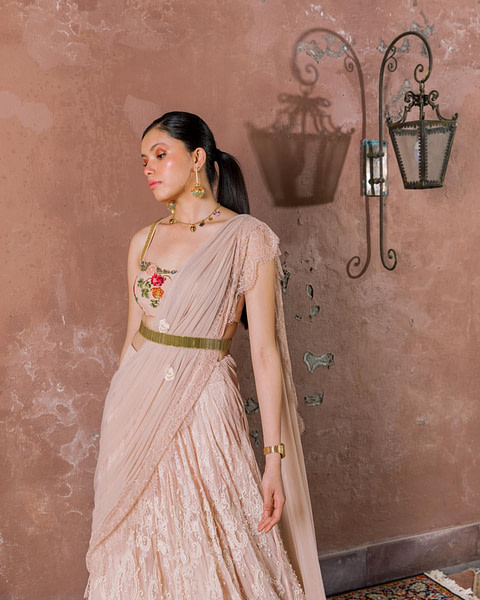 A woman in a beige embroidered saree with a rose-adorned blouse gazes away, her elegant stance highlighted by vintage wall sconces.
