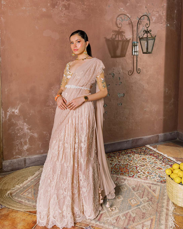 A woman in an ornate rose lace saree with pearl accents stands serenely, an orange belt adding a pop of color to the soft, romantic attire.