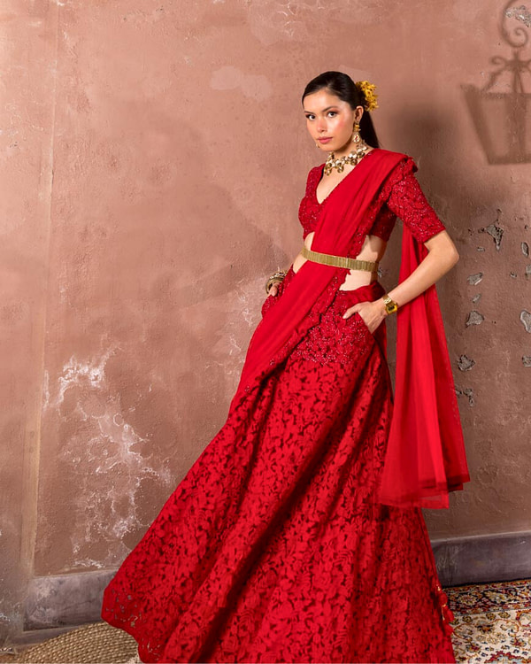 A confident woman stands against a textured wall in a lavish red dress with intricate embroidery, embodying a blend of tradition and modern style.