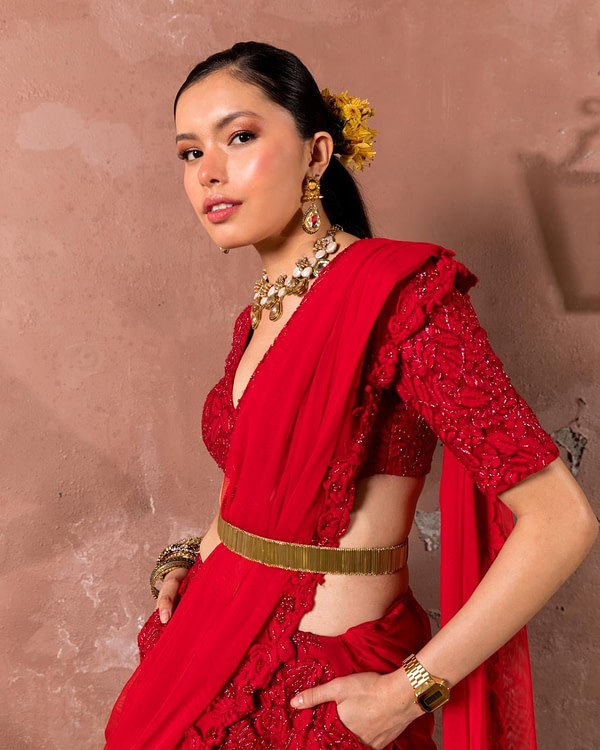 An elegant woman poses in a red embroidered outfit with a deep neckline, accessorized with a bold golden necklace, earrings, and floral hair decor.