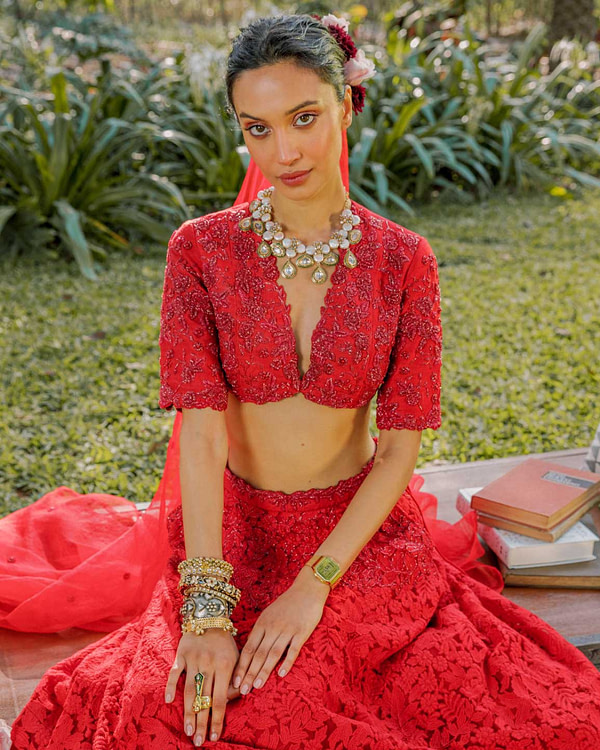 Close-up of a woman in a red lace lehenga, featuring a deep neckline and intricate patterns, complemented by a bold necklace, bangles, and a floral hairpiece.
