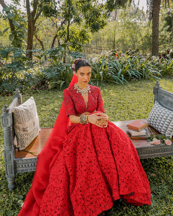 A seated woman poses in a garden, draped in a red lace lehenga and dupatta, with a backdrop of green foliage, accessorized with layered bracelets and a statement necklace.