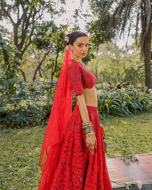 A woman stands in a lush garden, wearing a red lace embroidered lehenga with a matching cropped blouse and sheer dupatta, adorned with a floral hair accessory and statement jewelry.