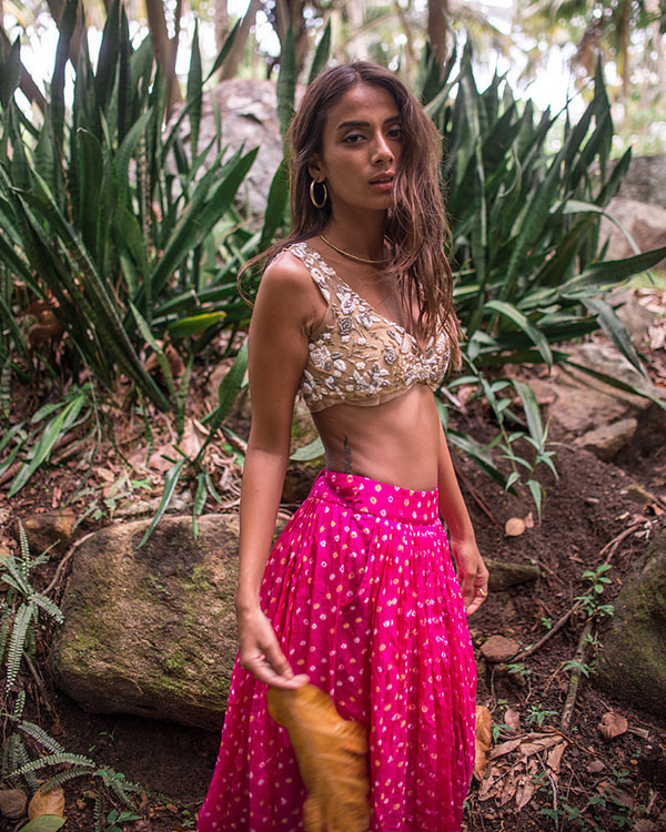 Captured from behind, a woman glances over her shoulder, her nude embroidered blouse contrasting with the lush greenery and pink bandhani skirt.