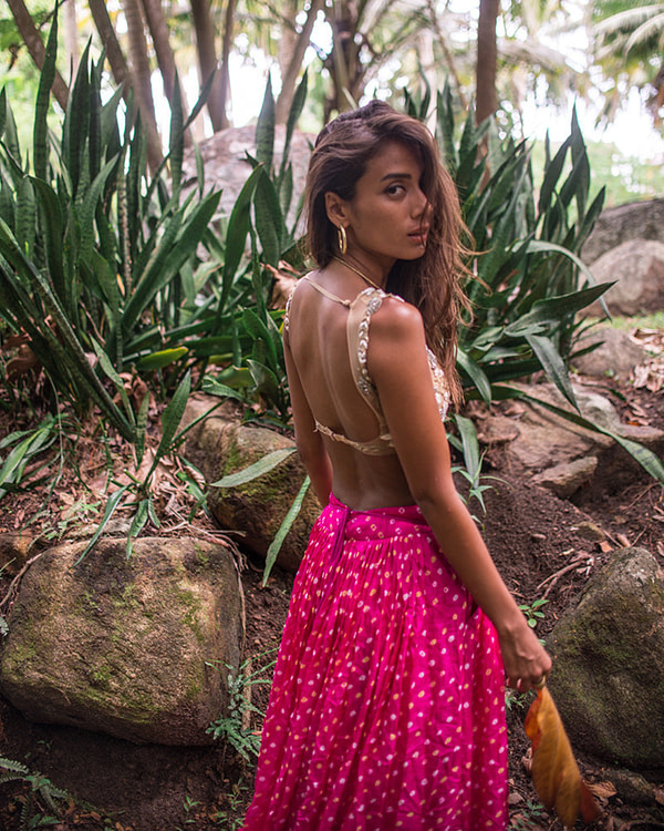 A woman in a tropical setting wears a nude embellished blouse and vibrant pink bandhani skirt, evoking a sense of bold, carefree elegance.