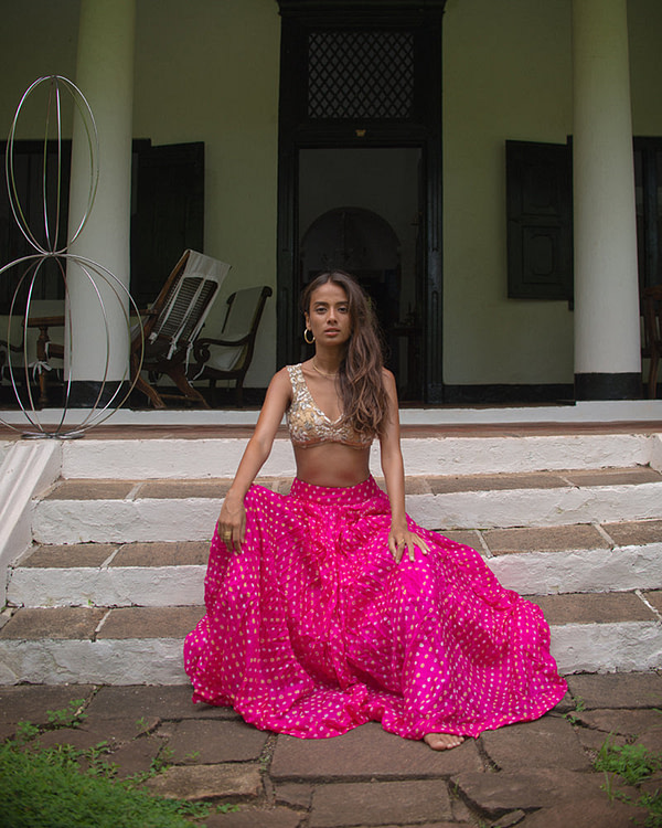 A woman sits on steps outside a colonial-style building, her attire a blend of tradition and modernity with a beaded blouse and bright pink skirt.