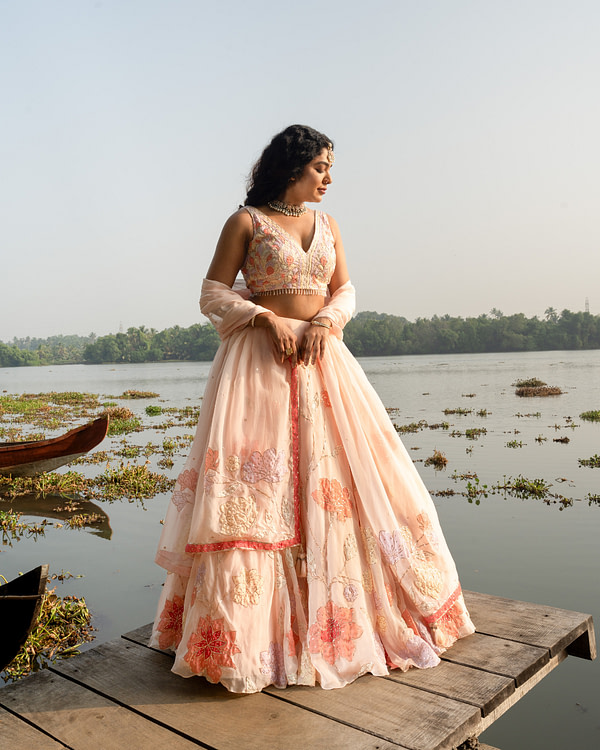 Bride on a pier in a blush lehenga with floral embroidery, looking to the side, with a serene lake and lush trees behind her.