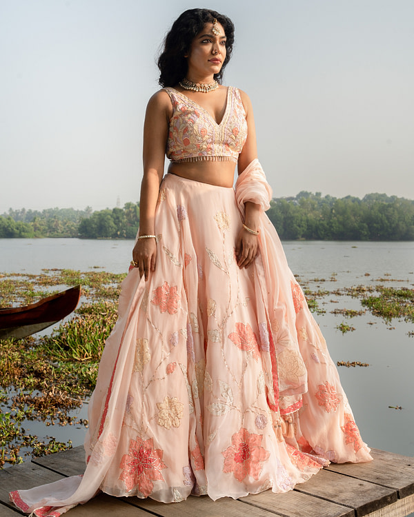 Elegant bride standing on a wooden pier in a blush embroidered lehenga, with a serene lake and greenery in the background.